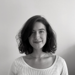 A person wearing a white shirt with shoulder length brown hair standing in front of a grey wall.