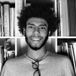 A person smiling with glasses hanging from their shirt while sitting in front of a bookshelf.