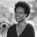 A woman who is speaking and sitting in front of a vase full of sunflowers.