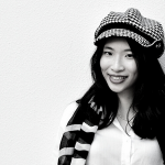 A person with a black and white hat and scarf smiling in front of a white wall.